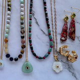 A collection of jewelry laid out on a white marble surface, including gold chain necklaces, beaded necklaces in green, brown, and red, a white flower pendant, red patterned earrings, and gold hoop earrings.