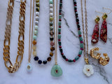 A collection of jewelry laid out on a white marble surface, including gold chain necklaces, beaded necklaces with green, red, brown, and white beads, gold hoop earrings, and a pair of red patterned drop earrings with gold fishhook wires.