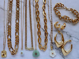 A collection of gold-toned jewelry including multiple necklaces, bracelets, and hoop earrings arranged vertically on a white marble surface. The set includes chunky and delicate chains, a turquoise pendant, a white flower charm, and matching gold hoop and stud earrings.