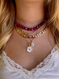 A close-up of a person's neck and chest wearing a layered necklace set. The set includes a magenta bead choker, a gold chain layer, a gold Figaro chain layer, and a delicate gold chain with a white flower and teardrop charm.